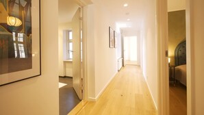 A hallway with wooden flooring, white walls, and a door leading to a bedroom with a bed and a nightstand.