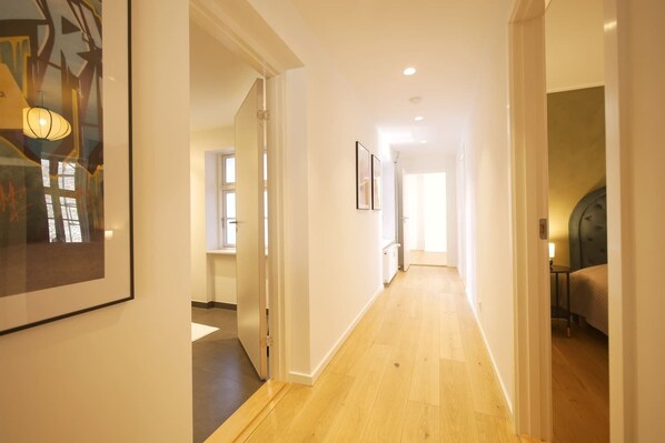 A hallway with wooden flooring, white walls, and a door leading to a bedroom with a bed and a nightstand.