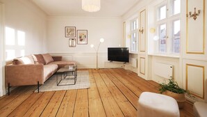 A living room with a sofa, a coffee table, a TV, and a large window with a view of buildings. - New Harbour / Carolina's Apartments / 2 BR (København)