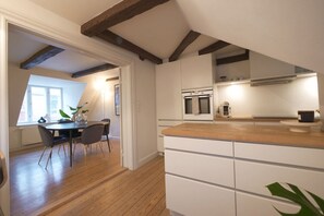 A modern kitchen with white cabinetry and a wooden countertop, a dining area with a table and chairs, and a view of the outdoors through a window.
