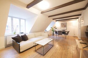 Eine moderne Loft-Wohnung mit geneigter Decke, einem weißen Sofa, einem Glastisch, einem Essbereich und einem gro ßen Fenster, das für natürliches Licht sorgt.