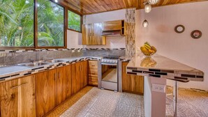 A modern kitchen with wooden cabinets, a central island, and a view of palm trees through large windows. - lazy parrots' luxury apartment#1: ✔pool ✔beach path✔AC (Puerto Viejo de Talamanca)