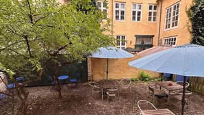 An outdoor patio with a brick floor, metal chairs, and tables. There are umbrellas providing shade.
