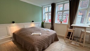 A neatly made bed with a quilted cover, a small table with a chair, and a window with curtains.