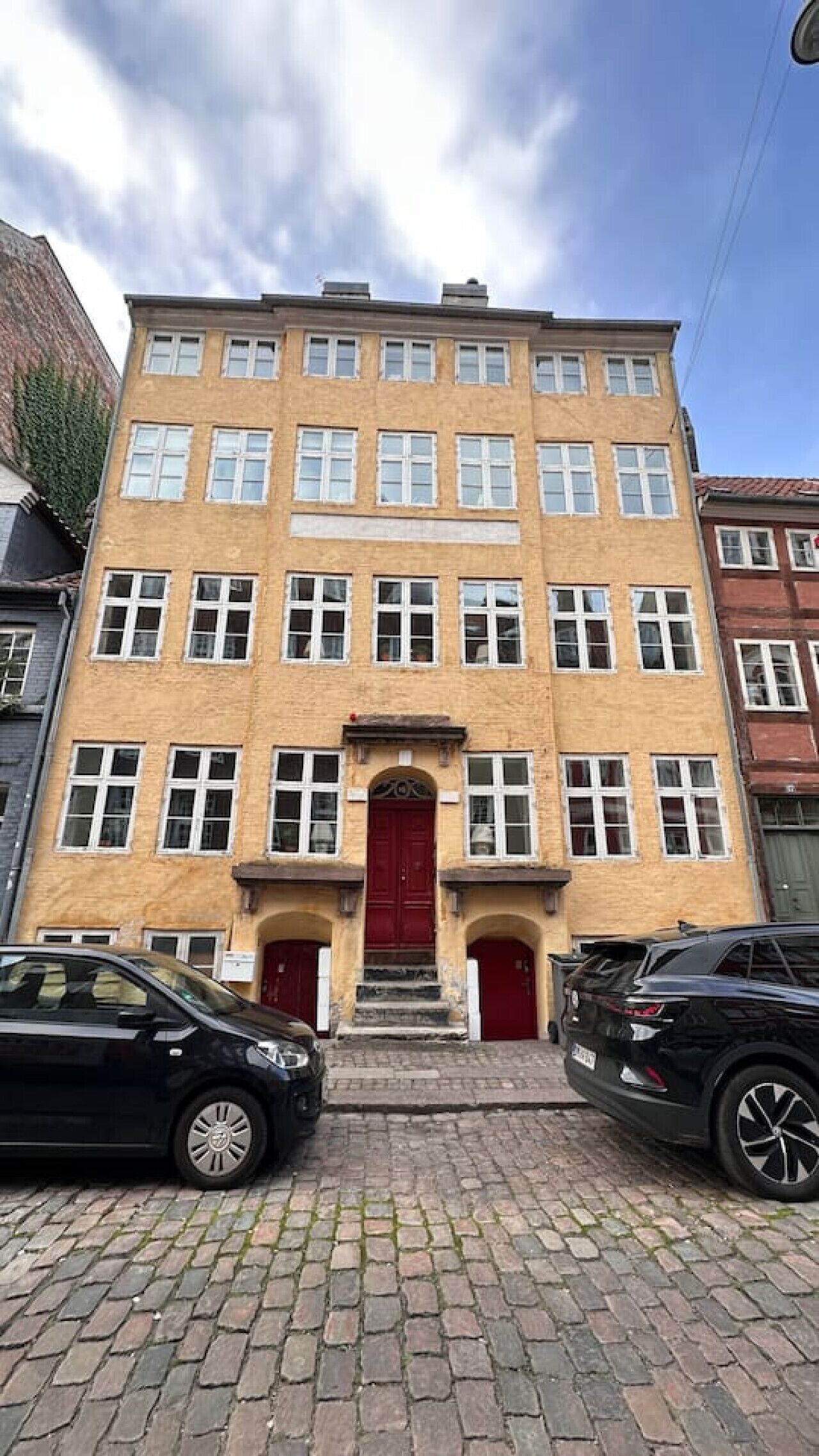 Um edifício de vários andares com uma porta vermelha central, várias janelas e fachada de tijolos.