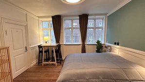 Een slaapkamer met een bed, een bureau en twee stoelen. Er is een raam met gordijnen, een plant en een lamp op het bureau.