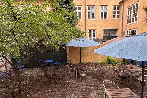 An outdoor patio with a brick floor, wooden tables and chairs, and blue umbrellas providing shade.