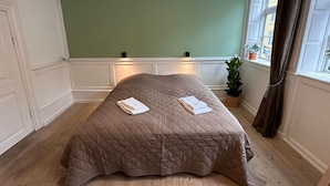 Une chambre avec un grand lit, des murs verts, une fenêtre avec des rideaux et une plante en pot.