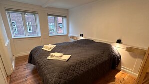 Ein Schlafzimmer mit einem großen Bett, zwei Fenstern, Holzboden und einem kleinen Nachttisch.