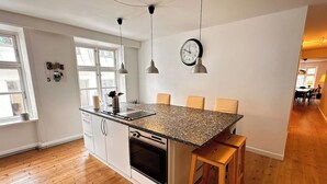 A modern kitchen with a granite countertop, white cabinetry, and a built-in oven. There are two pendant lights, a wall clock, and a window with blinds. - Main shopping street RAP 7 (København)