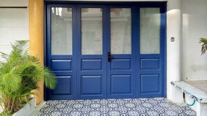 A blue double door with a textured glass panel, a potted plant to the left, and patterned floor tiles. - Casa Colors (Cartagena)