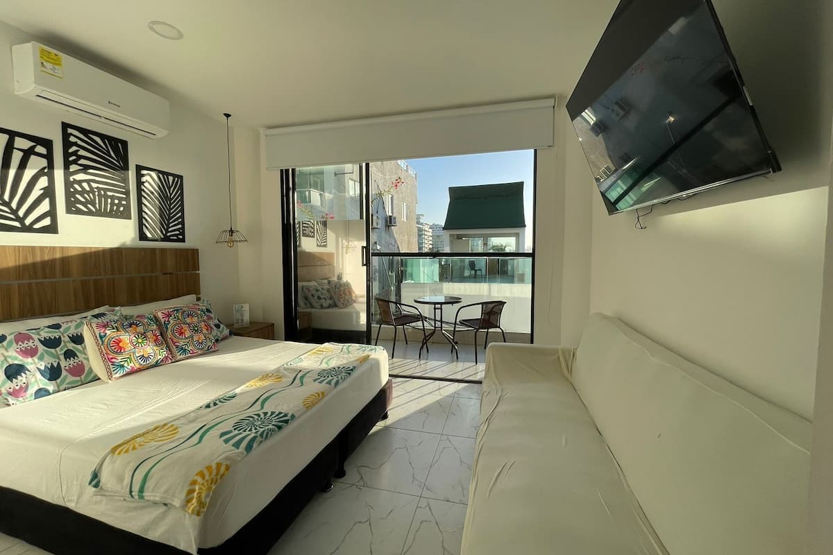 A modern hotel room with a large bed, a sofa, a balcony with a table and chairs, and a TV mounted on the wall.