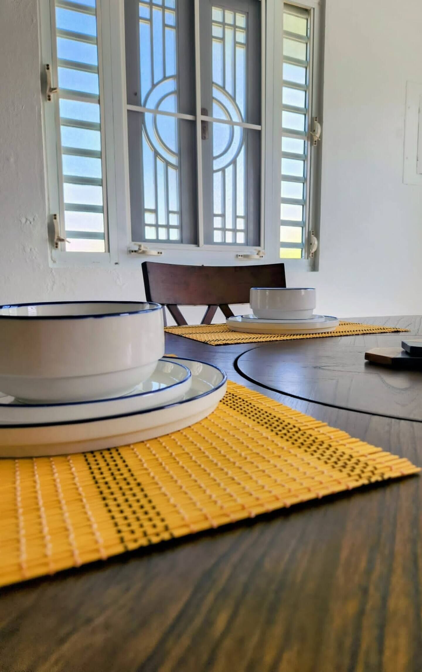 Un coin repas avec une table dressée pour deux personnes, agrémentée d’assiettes blanches à bordure bleue, d’un bol et d’une tasse. La table est posée sur un plancher en bois, et des sets de table jaunes tissés y sont disposés.