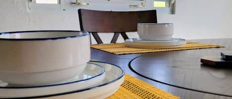 A dining area with a table set for two, featuring white plates with blue rims, a bowl, and a cup. The table is on a wooden floor with yellow woven placemats.