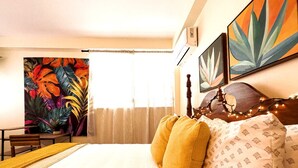 A hotel room with a bed, a wooden headboard, a painting of tropical leaves, and a window with curtains. - Two floor combo unit (San Juan)