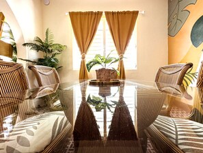 A room with a glass table surrounded by wicker chairs, a potted plant, and a large window with curtains.