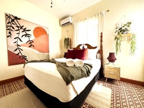 A neatly made bed with a blanket and rolled towels, a bedside plant, and a wall-mounted air conditioner.