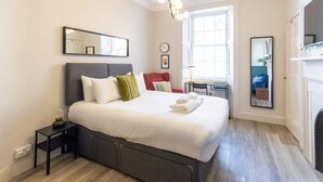 Un dormitorio con cama, mesita de noche, lámpara, silla y ventana.