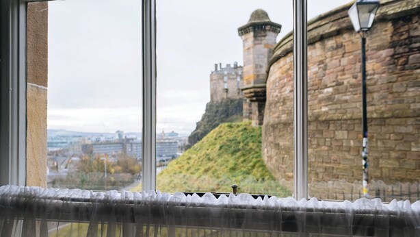 Vista a través de una ventana con cortina, que muestra un muro de piedra y una estructura similar a un castillo al fondo.