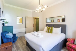 A hotel room with a bed, a blue chair, a small table, a mirror, and a green door.