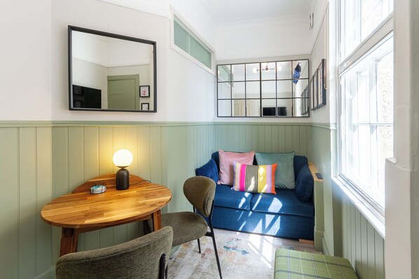 A room with a wooden table, a chair, a blue sofa with colorful pillows, and a mirror on the wall.