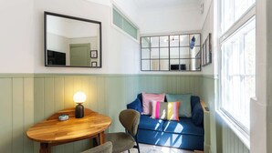Una habitación con una mesa de madera, un sofá azul con cojines coloridos, un espejo y una ventana con luz natural.
