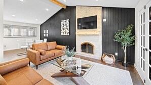 Een moderne woonkamer met een bruine leren bank, een glazen salontafel, een open haard en een grote tv aan de wand.