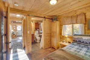 Ein Holzhütten-Innenraum mit einem Bett, einem Nachttisch, einem Fenster mit Vorhängen und einem durchgegangenen Türspalt erkennbaren Badezimmer.