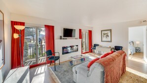 Uma sala de estar moderna com sofá, mesa de centro e TV. Possui uma varanda com cortinas vermelhas e vista para o exterior.