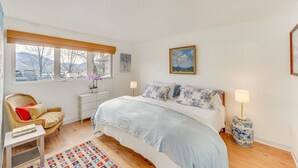 Um quarto com cama, uma cadeira, mesa de cabeceira, abajur, janela e um quadro na parede.