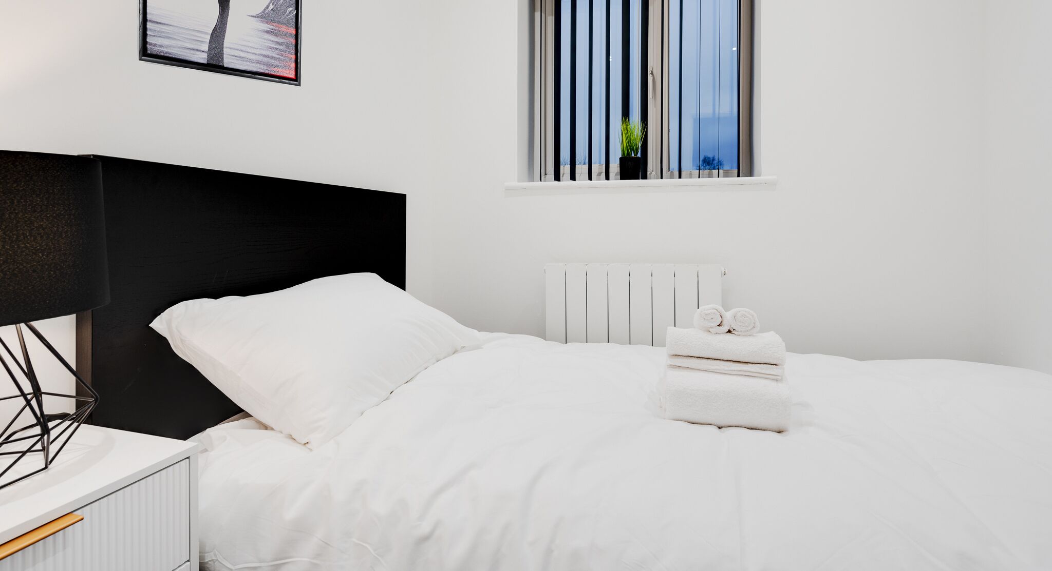 A modern bedroom with a black headboard, white bedding, a window with blinds, and a framed picture on the wall.