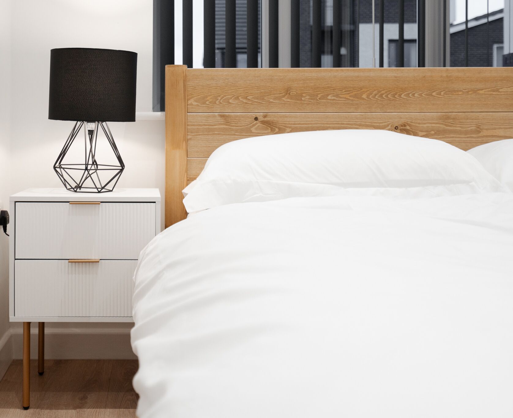 A bedroom with a wooden headboard, white bedding, a bedside table with a geometric lamp, and a white nightstand.