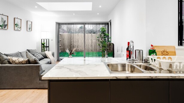 Una cocina moderna con encimera de mármol, un sofá gris y un amplio ventanal corredero que da a un espacio exterior verde.