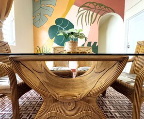 A dining area with a woven table and chairs, a potted plant, and a colorful wall mural.