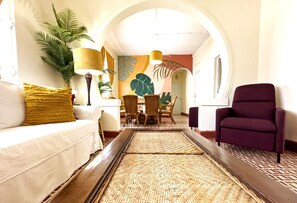 A hotel lobby with a white sofa, a purple armchair, a dining area with wicker chairs, a large plant, and a wall mural.