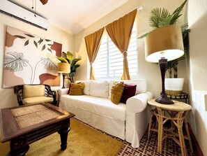A living room with a white sofa, a wooden coffee table, a lamp, and a large window with curtains.