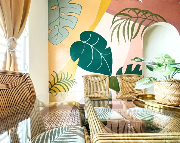 A room with wicker furniture, large tropical leaf wall art, and a potted plant.