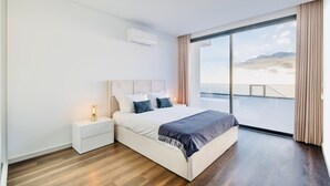 A modern bedroom with a large bed, a nightstand, a bedside lamp, and a view of the sky through a large window. - Vila Cales e Chadas by Your Madeira Rentals (Arco da Calheta)