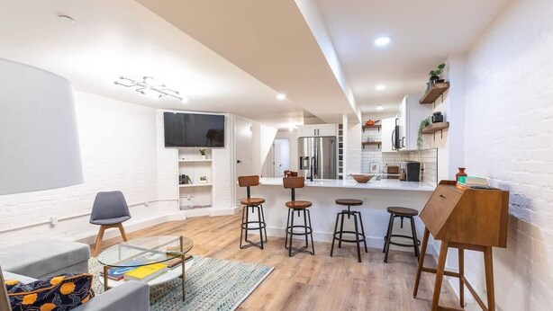 A modern living room with a glass coffee table, a sofa, and a TV. There are three bar stools and a wooden desk. The room has wooden flooring and white walls. - Sojourn in DuPont Circle: Modern Meets Historic (Washington)