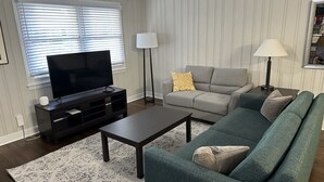 Ein Wohnzimmer mit einem Flachbildfernseher auf einem Lowboard, einem grauen Sofa, einem grünen Sofa, zwei Stehlampen und einem gemusterten Teppich.