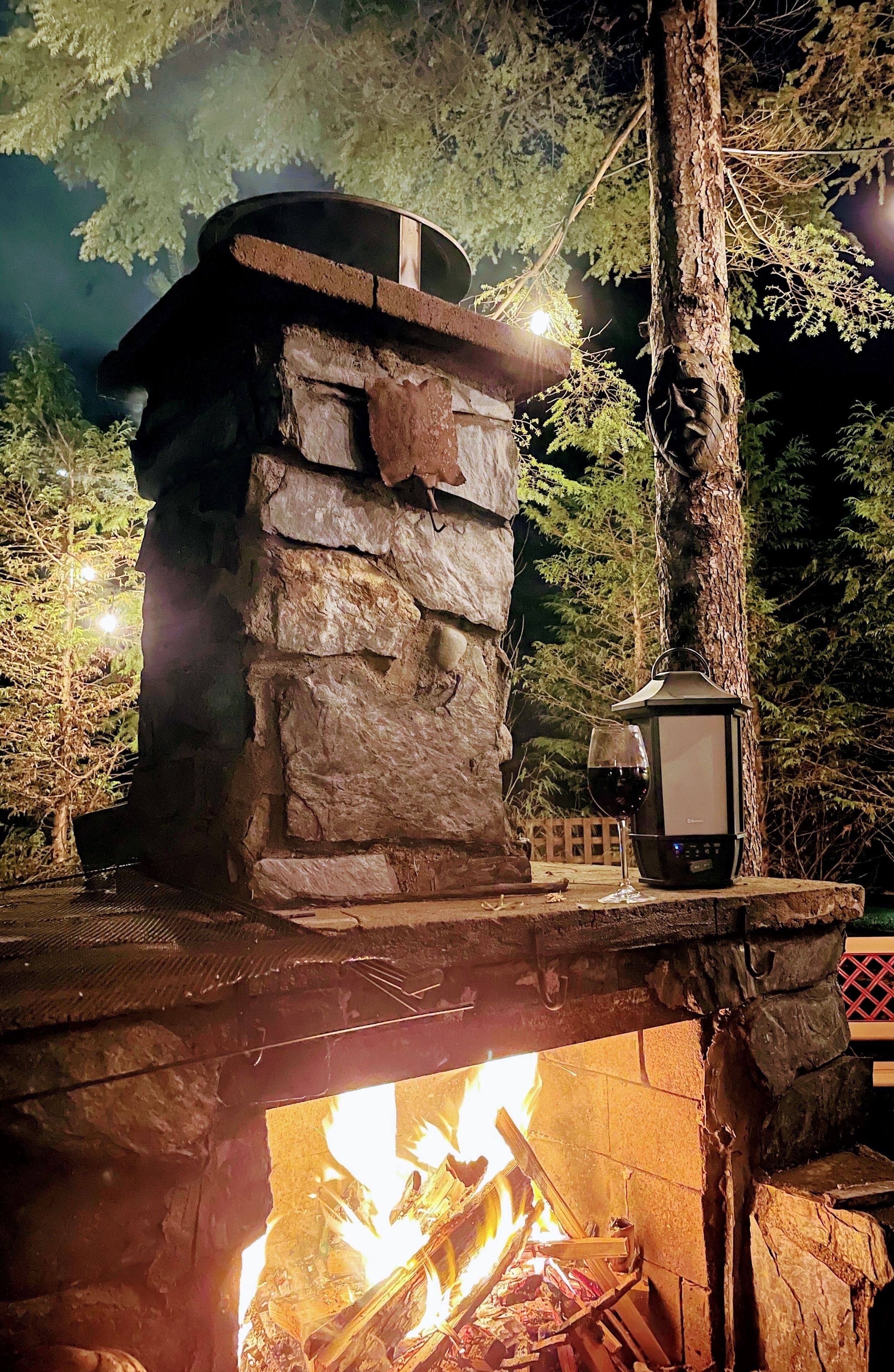 Una chimenea de piedra al aire libre con un farol y una copa de vino tinto, rodeada de árboles.