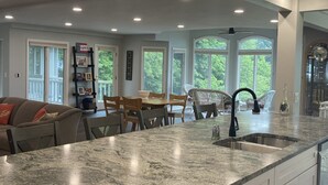A modern kitchen with a marble countertop, a dining area with a table and chairs, and large windows offering a view of greenery. - Spacious multi-level lakeside home w/ renovated kitchen and abundant parking!  (Penn Yan)