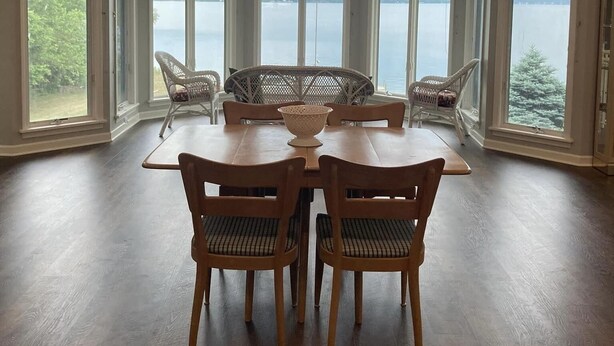 A dining area with a view of a lake, featuring a table with chairs, a ceiling fan, and large windows. - Spacious multi-level lakeside home w/ renovated kitchen and abundant parking!  (Penn Yan)