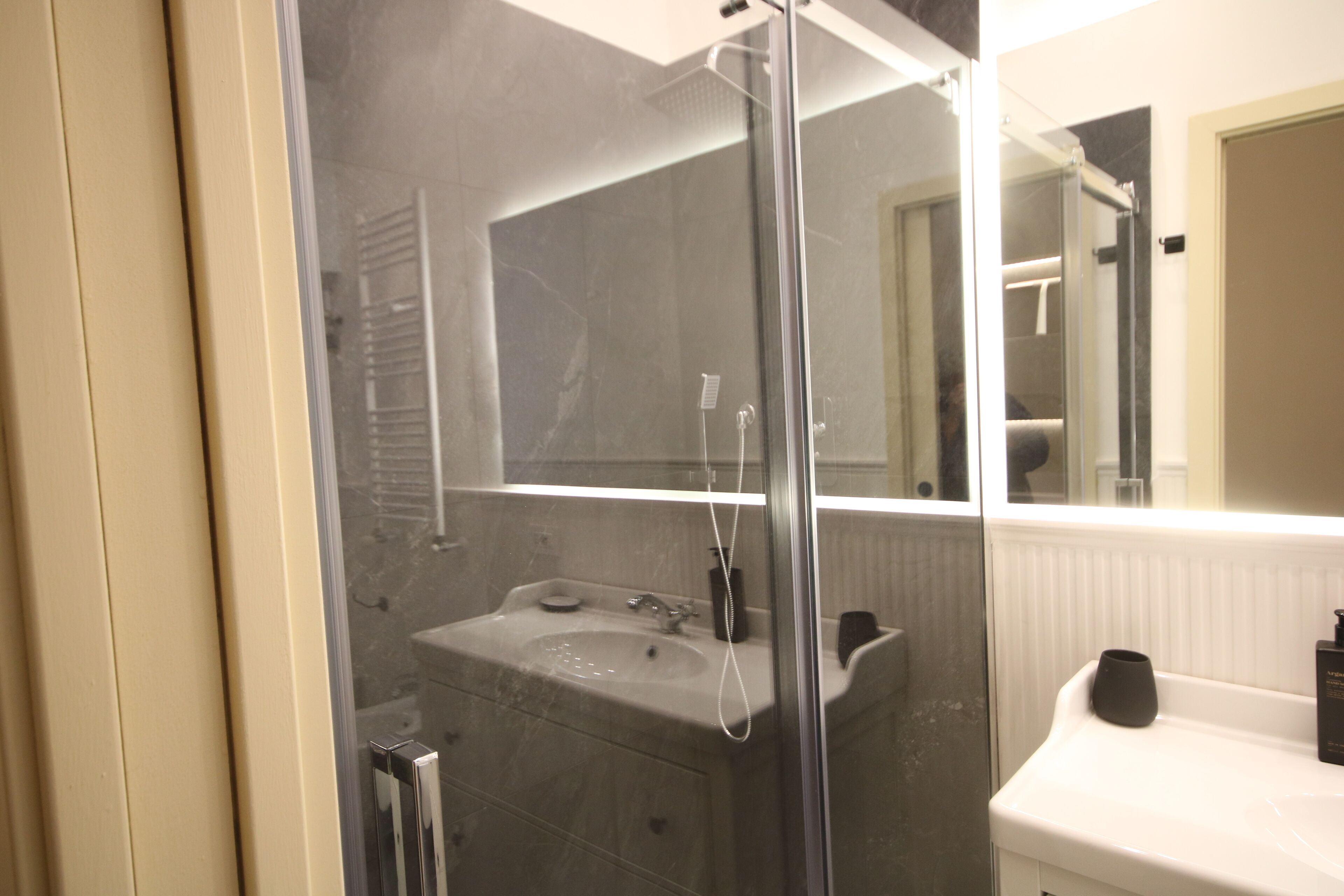 Un baño con ducha de vidrio, un lavabo con espejo y una encimera blanca con un objeto negro encima.