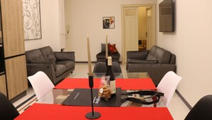 Un salon moderne avec une table à manger à plateau en verre, deux canapés gris et un téléviseur à écran plat fixé au mur.