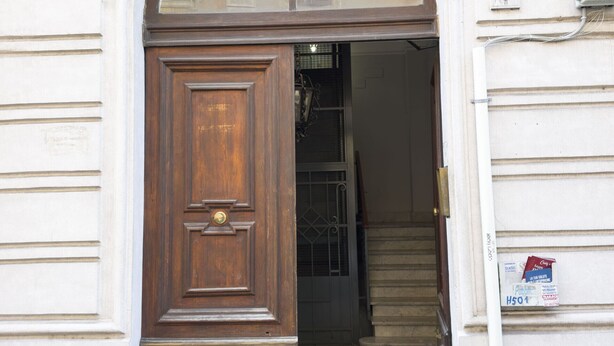 Une porte en bois, dotée d’une poignée en laiton et d’une plaque métallique, est intégrée à un bâtiment blanc dont la base est en pierre et au-dessus de laquelle se trouve une petite fenêtre.