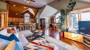 A spacious living area with a high ceiling, wooden beams, a large window, and a television. - The Kebler Home: Walk to Elk Ave! Dog Friendly! (Crested Butte)