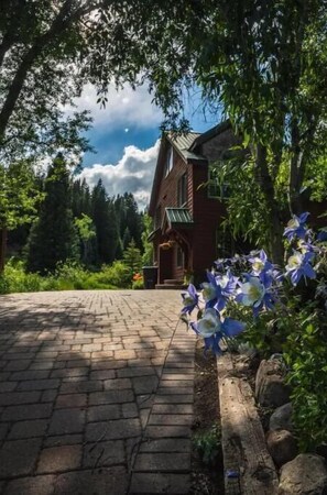 Une maison de campagne au charme rustique, avec un chemin de briques menant à l’entrée, entourée d’une végétation luxuriante et d’arbres.
