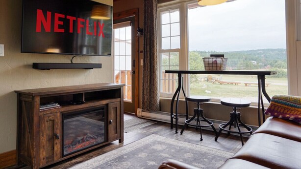 Ein Wohnzimmer mit an der Wand montiertem Fernseher, einem Holzschrank, einem Tisch mit Hockern und Blick ins Freie.