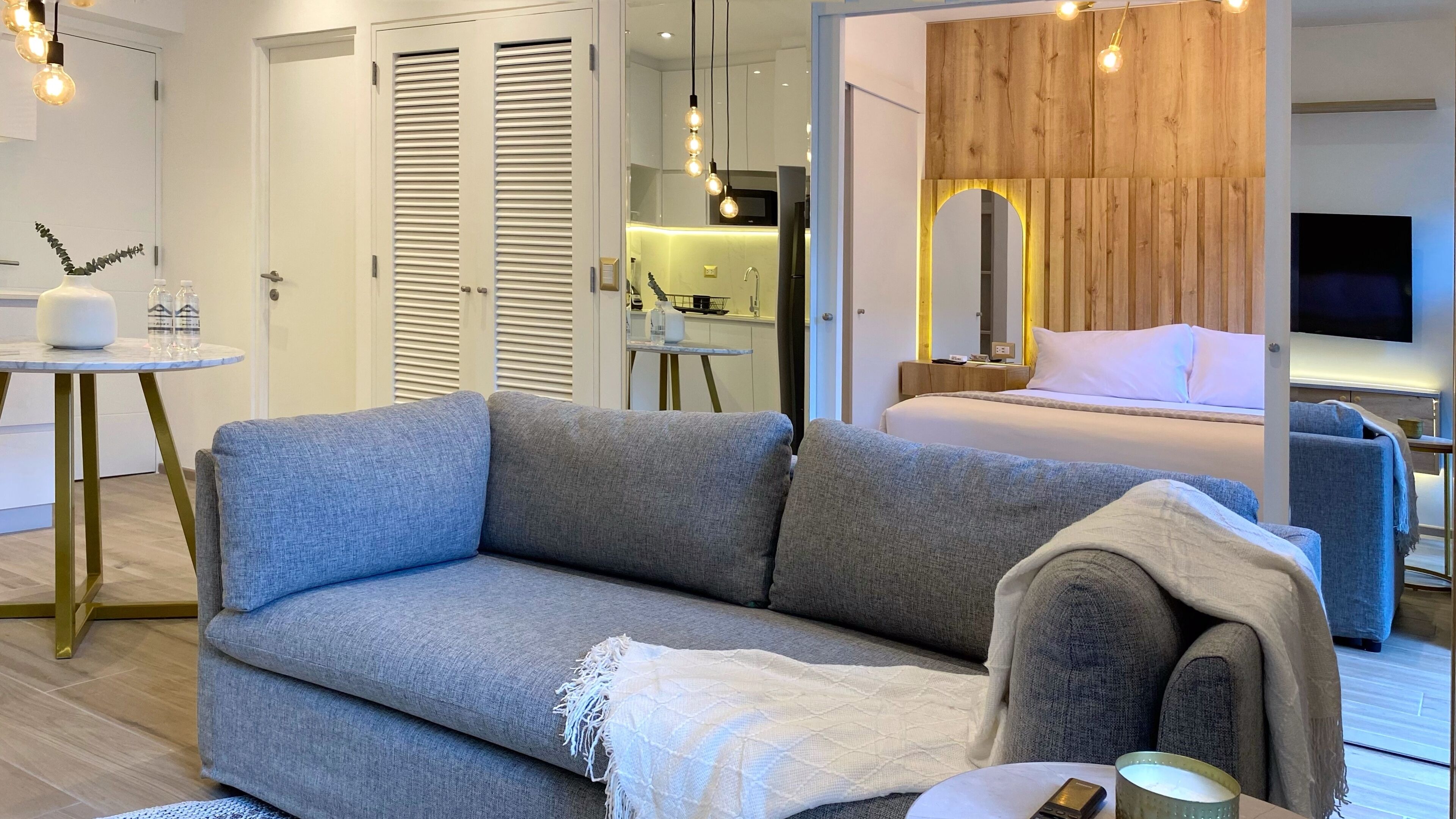 Una habitación de hotel moderna con un sofá gris, una mesita redonda pequeña, una cama con cabecera de madera y un televisor montado en la pared.